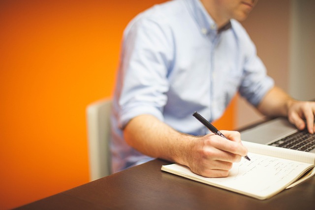 A man taking notes in a notebook while using a laptop