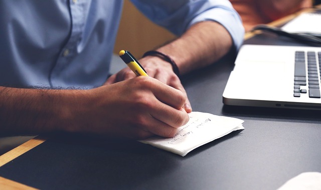 A person writing notes on paper next to a laptop