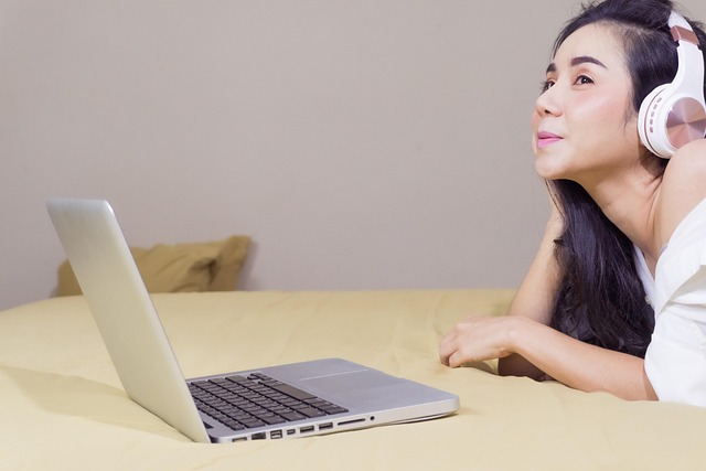 A woman with headphones looking at a laptop on a bed